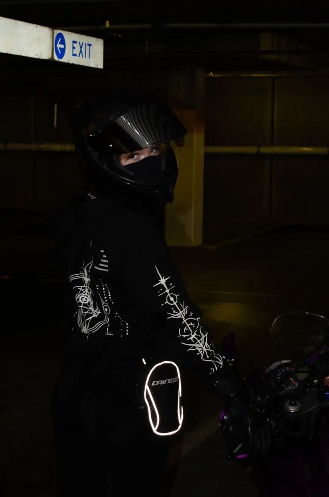Rider in a full-face helmet turned to the side in a dark garage, the COMMIT hoodie sleeve art and back print glowing