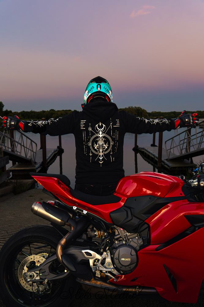 Rider with arms spread behind a red Ducati at sunset, showing the COMMIT Hoodie Proto back print and both sleeves
