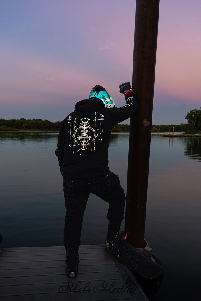 Rider standing on a dock at sunset with their back to the camera, the COMMIT Hoodie Proto sigil back print in full view