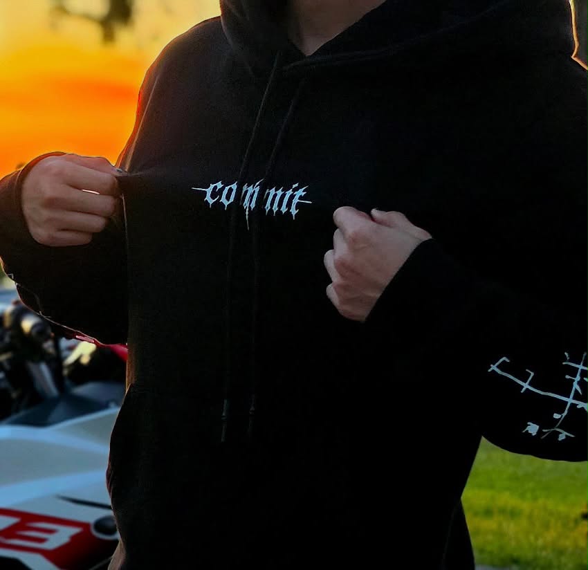 Hands gripping the front of a black COMMIT hoodie at sunset, chest logo and sleeve sigil in view